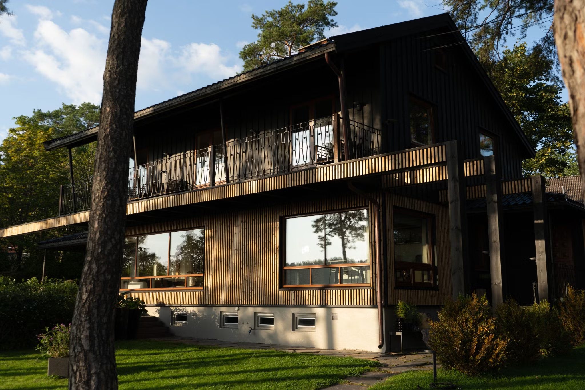 Modernes zweistöckiges Haus mit großen Fenstern und Aluwood-Fassadenpaneelen aus dunklem Holz, umgeben von Bäumen und grünem Rasen unter Sonnenlicht.